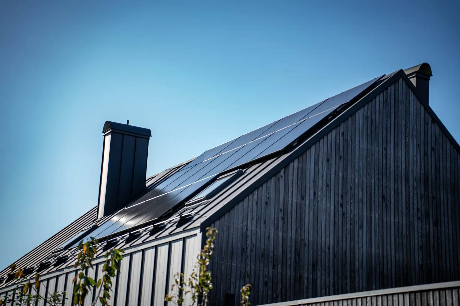 Helsvarte solcellepaneler på taket av et moderne hus med mørk trekledning, mot blå himmel